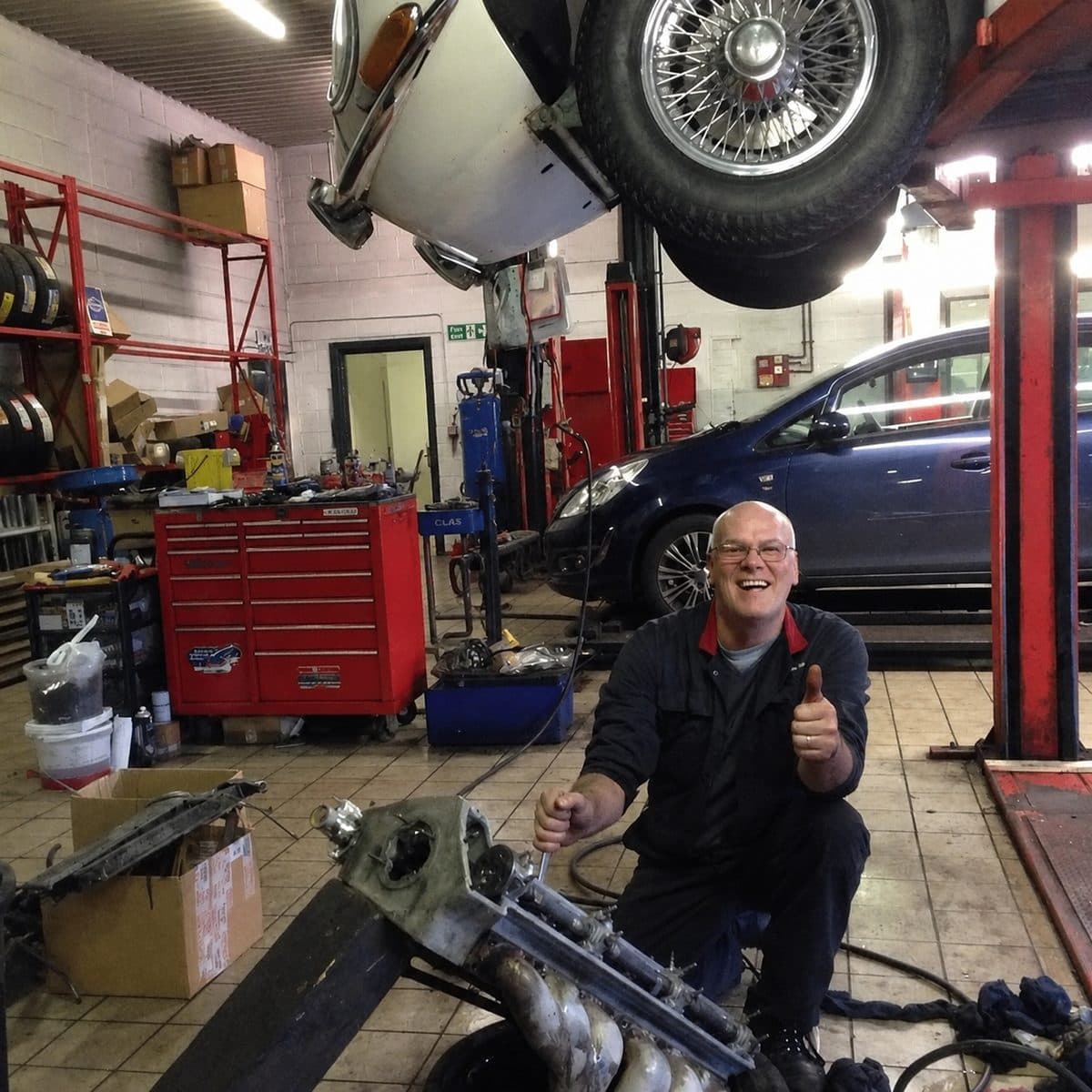 Keith Wheeler crouched on the workshop floor in Dudley giving a thumbs up, with an exhaust manifold in front of him and a car raised on the lift behind
